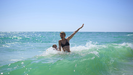 Summer happy family of six years blonde child playing and jumping water waves embracing woman mother in sea shore beach