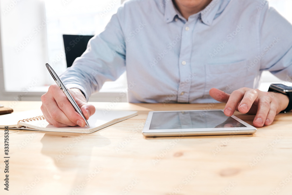 Aufkleber Close-up of unrecognizable businessman sitting at desk and using tablet while making notes about promotion statistics