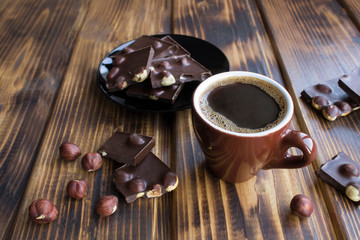 Black  coffee in the brown ceramic cup and pieces of dark chocolate with hazelnuts on the brown wooden  background. Closeup.