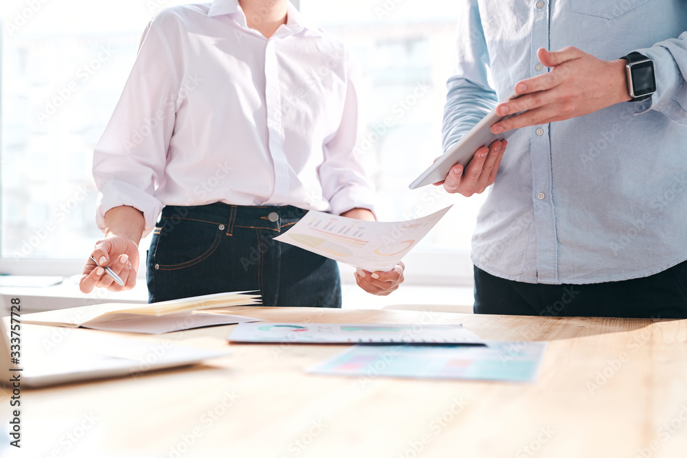 Poster close-up of unrecognizable colleagues standing at table and discussing financial papers in office