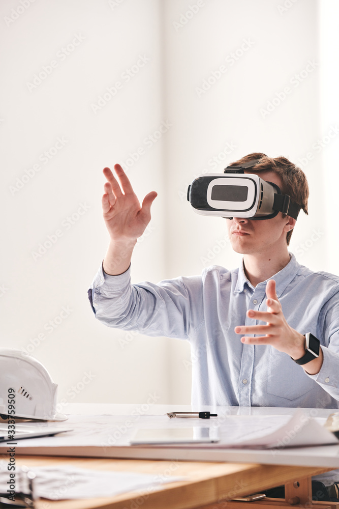 Aufkleber Male engineer sitting at desk and gesturing hands while designing 3D model of building with VR device