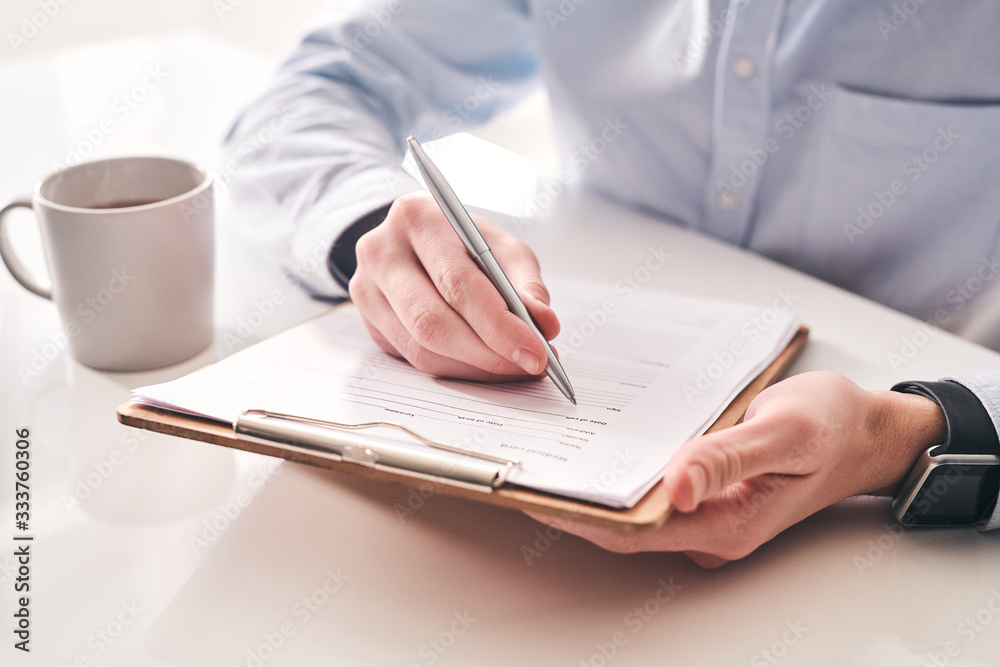 Poster Close-up of unrecognizable businessman sitting at table with tea mug and making notes in application form