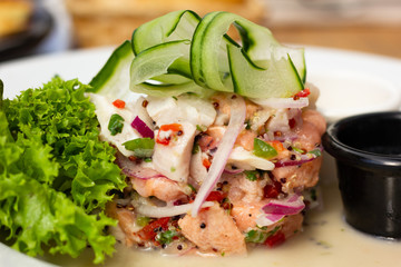 Fresh mixed seafood ceviche plate with zucchini on top and lettuce on side. Chopped salmon, prawns, tomato, peppers and onion slices on white plate. Gourmet restaurant food concept