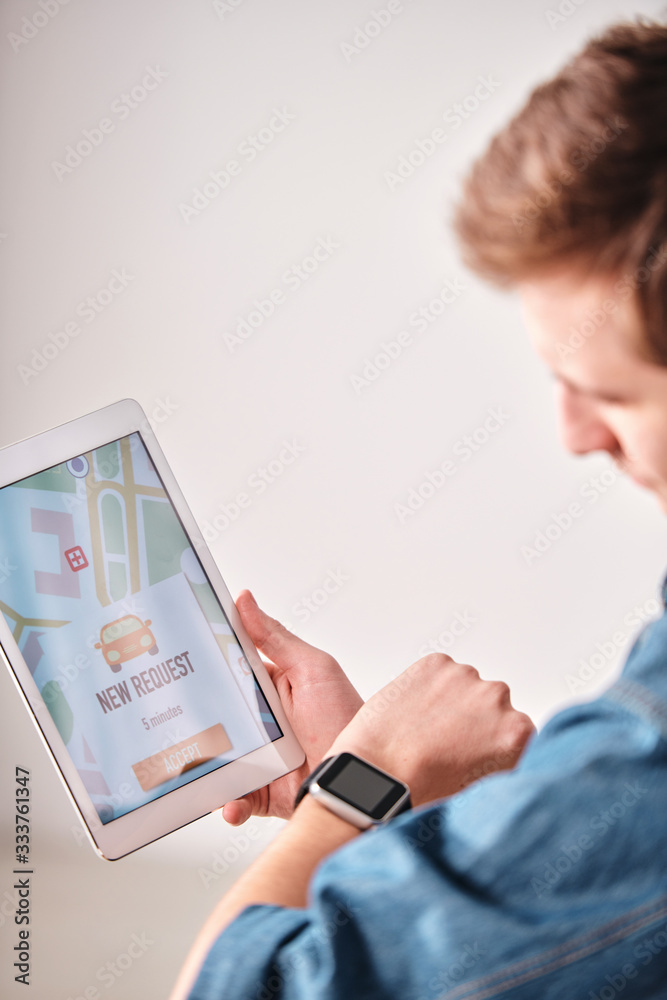 Poster Over shoulder view of young man checking time on smart watch while ordering taxi online via tablet app