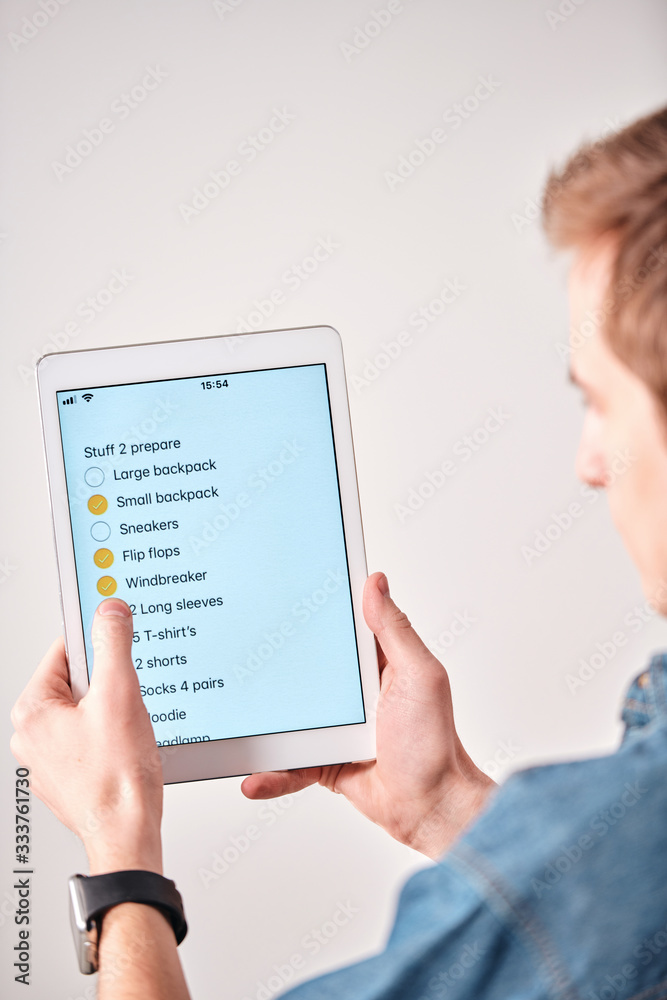 Poster Over shoulder view of young man using packing list in tablet note while putting checkmark in packed stuff