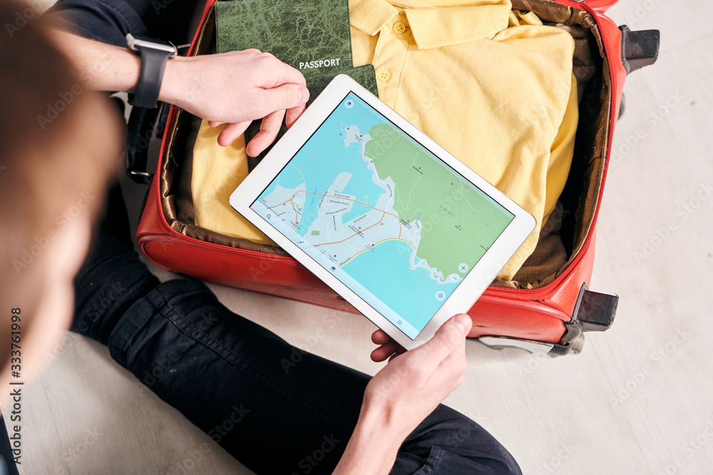 Poster Above view of unrecognizable man packing suitcase and using online map on tablet while planning tourists route in foreign country