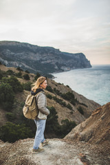 Naklejka na ściany i meble Happy young traveler woman walking and enjoying amazing coastline mountain view. With beige backpack, wearing in jacket and jeans.
