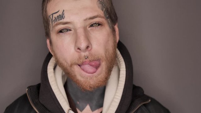 Man with tattoo on his face and neck, eyeball or eye scleral tattooing, piercing demonstrating splitted tongue (body modification), posing in studio. Isolated on grey background.
