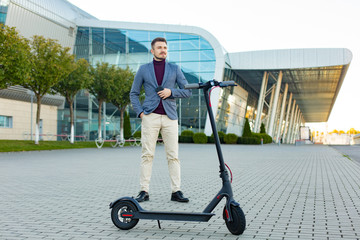 Young handsome stylish man with e-scooter standing on sidewalk near the airport on the sunset. Trendy urban transportation on modern electric scooter. Eco friendly mobility concept.