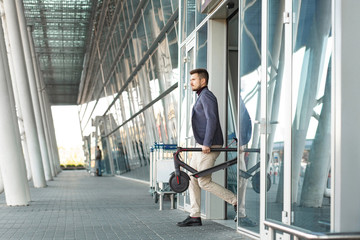 Fototapeta premium Young handsome man holding e-scooter leaves the airport. Rent a transport on a trip. Trendy urban transportation on modern electric scooter. Eco friendly mobility concept.