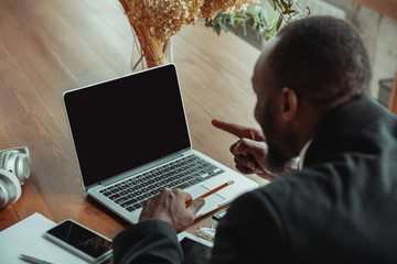 Businessman or student working from home being isolated or keep quarantine 'cause of coronavirus. African-american man using laptop, tablet and headphones. Online conference, lesson, remote office.