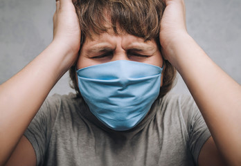 A young man in a blue protective medical mask respirator is holding head in hands on a gray wall...