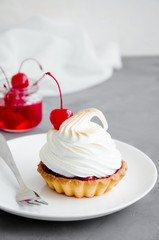 Tartalet with cherry filling and Italian meringue with a cocktail cherry on top on a white plate with a fork on a dark concrete background. Sweet food background. Cherry pie. Vertical, Copy space.