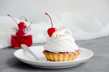 Tartalet with cherry filling and Italian meringue with a cocktail cherry on top on a white plate with a fork on a dark concrete background. Sweet food background. Cherry pie. Horizontal, Copy space.