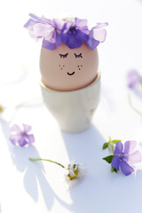 Obraz na płótnie Canvas Cute egg with face and flowers wreath on flowers background. Easter still life. Easter egg concept. Spring is coming. Minimal holiday concept. Cute creative photo. 