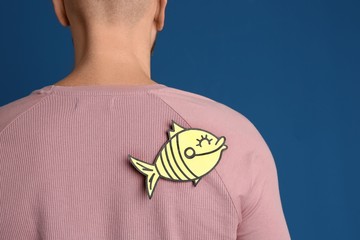 Man with paper fish on back against blue background, closeup. April fool's day
