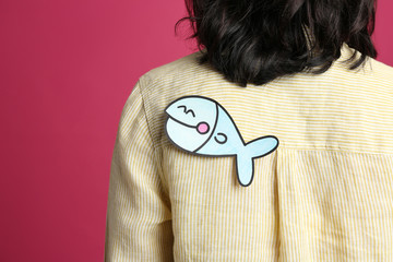 Woman with paper fish on back against pink background, closeup. April fool's day