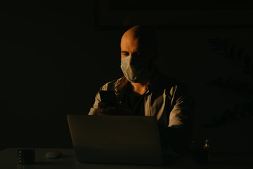 A man in a medical face mask working remotely during the quarantine to avoid the spread coronavirus. A businessman using the cellphone at home in the evening during the pandemic of COVID-19.