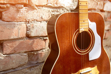 An old wooden guitar stands against a brick wall. The sun's rays fall on the guitar strings