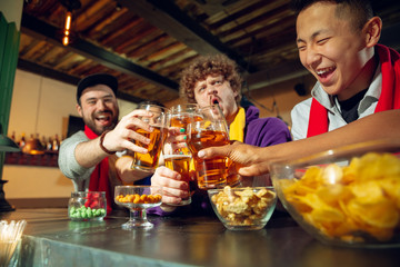 Sport fans cheering at bar, pub and drinking beer while championship, competition is going. Multiethnic group of friends excited watching translation. Human emotions, expression, supporting concept.