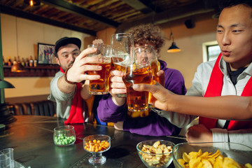 Sport fans cheering at bar, pub and drinking beer while championship, competition is going. Multiethnic group of friends excited watching translation. Human emotions, expression, supporting concept.