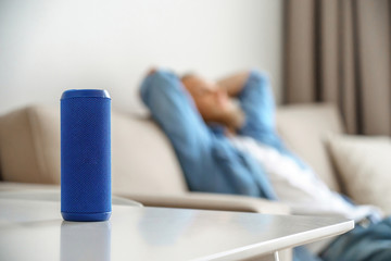 Small portable wireless speaker assistant on table with man lounge on sofa listening to music. Young relaxed guy enjoys mini bluetooth stereo gadget for sound digital assistance at smart home concept.