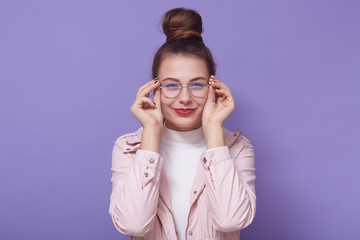 Studio portrait of woman with red lips, wearing stylish glasses, pink jacket and white, playfully looking at camera and tounching frame of her glasses, standing isolated over lilac studio background.