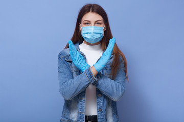 Horizontal shot of young beautiful woman dresses disposable flumask and rubber gloves doing stop sign, female standing isolated over blue background. Coronavirus, covid 19, heallth care concept.