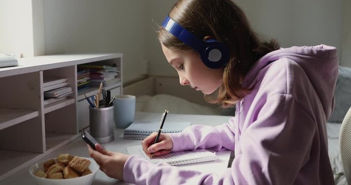 Focused teenage girl wearing headphones looking at smartphone using distance learning mobile education app. Teen school student studying online listening audio course making notes in copybook at home.