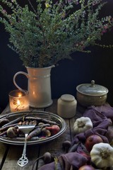Still life seen of root vegetables in a silver platter with a silver serving fork.  Jug of flowers and pots with a lit candle sit in the background.  Garlic bulbs and red onions sit to the right.