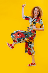 happy girl walk lifting a leg high, in colorful jumpsuit, holds a notepad and pencils in her hands, she has red curly hair, picture on a yellow background