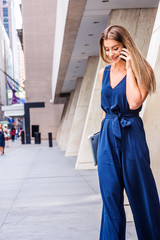 Young Eastern European American Woman talking on cell phone, traveling, working in New York City, wearing blue jumpsuit, carrying laptop computer, looking down,  walking out from office building..