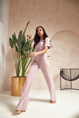 Beautiful sexy brunette woman tanned skin face cosmetic makeup wear pink suit pants for date walk office fashion clothes style collection interior room  sand color safari summer armchair palm boho.