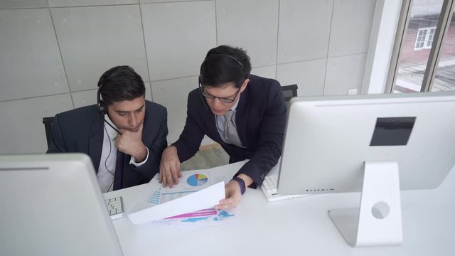 Professional Asian man customer support with colleague discussing business data to consulting and solving to customer. Male Broker telesales marketing teamwork working with using computer in office