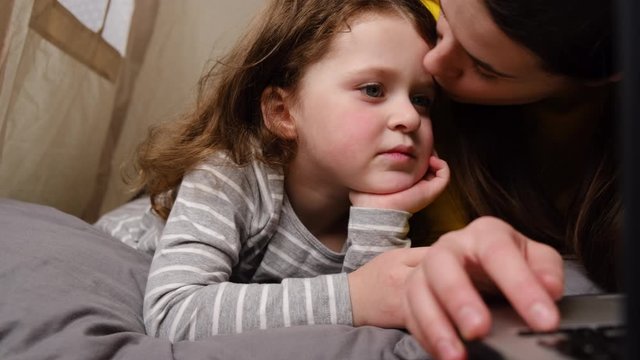 Smiling young mother and little child girl  having fun looking at laptop, sitting in tent, happy mom teaching  kid daughter use laptop application, watching cartoon or making video call on computer