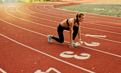 Young runner woman in sportwear getting ready to run sprint at low start on stadium track with red...