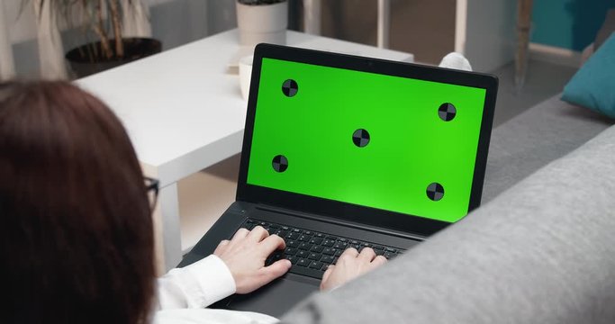 Close up of woman with brown hair working on laptop with green screen while lying on couch. Space for text and advertisement. Modern technology concept.