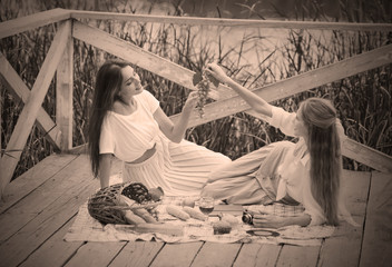 Fototapeta premium Two cheerful young women have a picnic outdoors on a summer day. Two girlfriends in retro vintage style clothes spend time together on the pond pier.