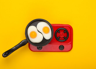 Miniature toy frying pan with fried eggs on stove. Yellow background. Top view. Minimalism. Studio shot