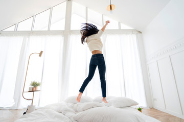 attractive asian Happy young woman in earphones is listening to music with smart phone, jumping on bed, singing and smiling white bedroom interior background freedom lifestyle activity
