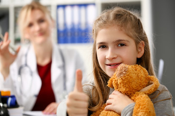Happy little child girl hold teddy bear and thumbs up in hospital female excel doctor office.