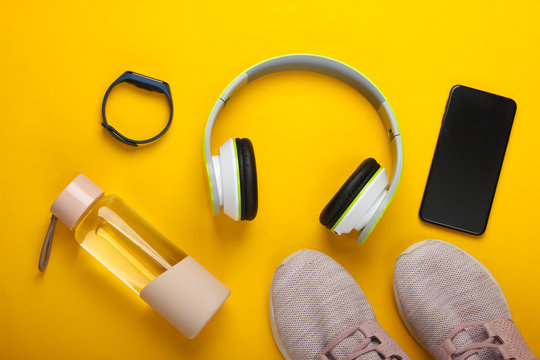 Flat lay composition of modern gadgets for sports and fitness. Wireless headphones, smart bracelet, smartphone, running shoes, water bottle on a yellow background. Top view