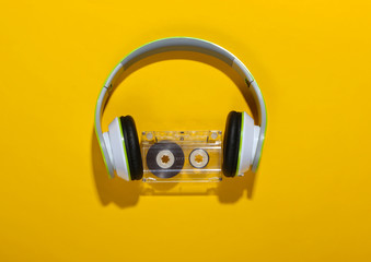 Stylish wireless stereo headphones and audio cassette on yellow background with shadow. Music lover. Retro 80s. Top view. - obrazy, fototapety, plakaty
