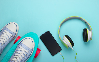Flat lay hipster composition. Sneakers on cruiser board and stereo headphones with smartphone on blue pastel background. Summer fun. Top view