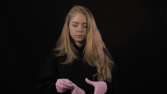 Young woman with pink gloves puts on a pink medical mask. Isolated on black background. Health care and medical concept. Close up portrait . 4k. Coronavirus Epidemic, illness, pandemic