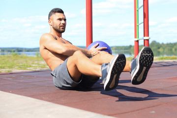 Image of handsome strong sports man doing exercises with fitness ball outdoors at sunny day