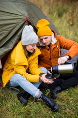 travel, trekking and hiking concept - portrait of couple sitting near green tent and drinking tea in autumn forest