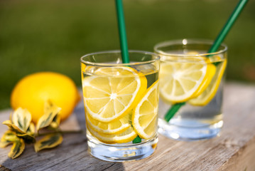 Summer drink, glasses of water filled with slices of lemons, stand on a wooden board on the lawn in sunny warm day, the background is blurred, shallow depth of field, selective focus.