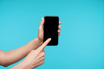 Close-up of black modern smartphone being kept by woman's hands with white manicure, touching screen with forefinger while standing over blue background