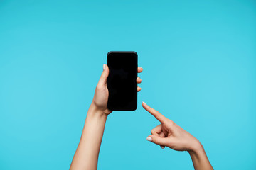 Horizontal shot of raised female's hands holding modern mobile phone and showing on black screen with index finger, isolated against blue background
